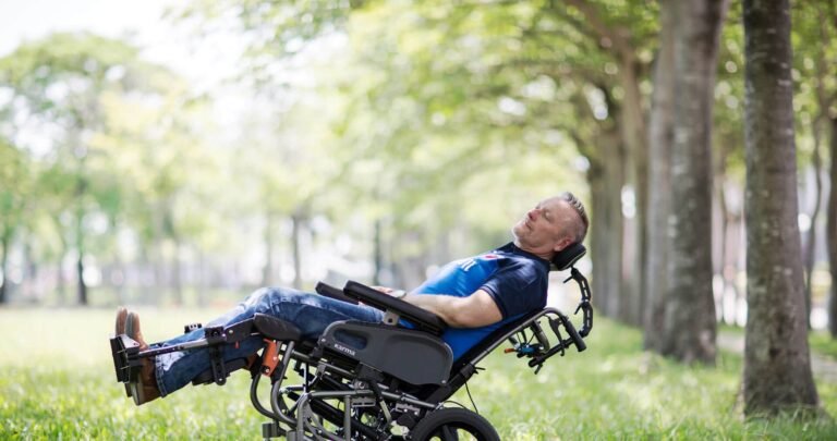 An elderly person sits in a comfortable modern wheelchair on a sunny garden patio.